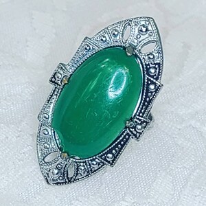 Puede incluir: Un anillo de tono plateado con una gran piedra cabujón ovalada de color verde, engastada en un marco de metal alargado e intrincado con recortes decorativos y pequeños acentos en forma de cuentas a lo largo de los bordes, creando un aspecto detallado.