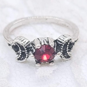 May include: A silver-toned ring featuring a large, round, red gemstone at the center. The ring has decorative side accents with black stones. The ring is set against a white background.