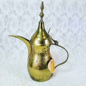 May include: A vintage brass coffee pot with a long spout, curved handle, and a decorative finial on the lid. The pot has a tarnished, aged appearance, and a small tag is attached. The background is a white, floral lace pattern.