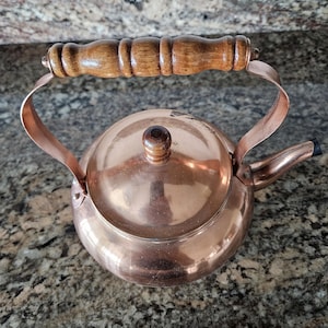 May include: A polished copper tea kettle featuring a rounded body, a short spout, and a removable lid with a small wooden knob. The kettle includes a curved metal handle topped with a textured, cylindrical wooden grip for handling.