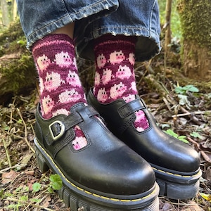May include: A pair of black leather T-strap shoes with thick lug soles and yellow stitching, worn with dark blue denim jeans and knit socks featuring a repeating pattern of pink and white pig faces on a deep magenta background.