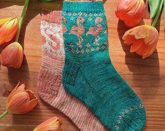 May include: A pair of knitted socks, one teal with a pink flamingo design and the other a peach color with a white design. The socks are lying on a wooden surface with orange tulips.