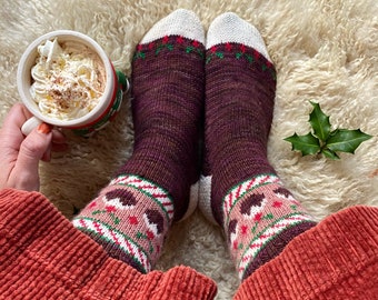 May include: A pair of brown and white knitted socks with a Christmas-themed design. The socks feature a candy cane stripe pattern with red and white accents, and a gingerbread man design with pink and brown details. The socks are worn by a person sitting on a white fluffy surface.