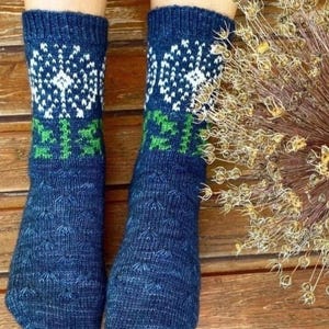 May include: A pair of dark blue knit socks featuring a white dandelion seed head pattern at the top, a green stem design below the flower, and a textured stitch pattern covering the lower foot area, crafted from a heathered blue yarn.