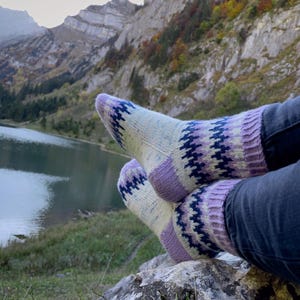 Könnte beinhalten: Gestrickte Socken mit einem geometrischen Muster in Creme, Lila und Marineblau. Die Socken werden mit dunkelblauen Jeans getragen. Im Hintergrund sind ein See und Berge zu sehen.