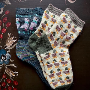 Könnte beinhalten: Zwei Paar gestrickte Socken. Ein Paar ist dunkelblau mit einem Muster aus lila, grauen und türkisfarbenen Tauben. Das andere Paar ist cremefarben mit einem Muster aus Enten in Lila-, Orange- und Grüntönen. Die Socken liegen auf einer dunkelbraunen Oberfläche.