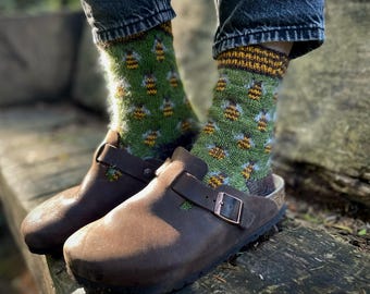 May include: A pair of brown leather clogs with green and yellow socks featuring a bee pattern. The socks are made of a soft, fuzzy material.