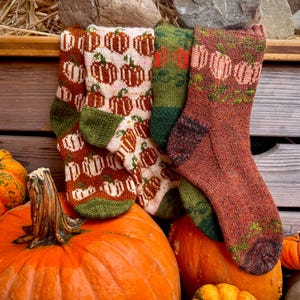 May include: Four knitted socks featuring pumpkin patterns. Two socks have a cream background with orange pumpkins and green cuffs, while the other two feature green and rust-colored yarn with pumpkin motifs and dark brown heels and toes.