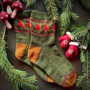May include: A pair of green knitted socks with a brown and red pattern of robins. The socks have a brown cuff and are lying on a dark brown surface with green pine branches.