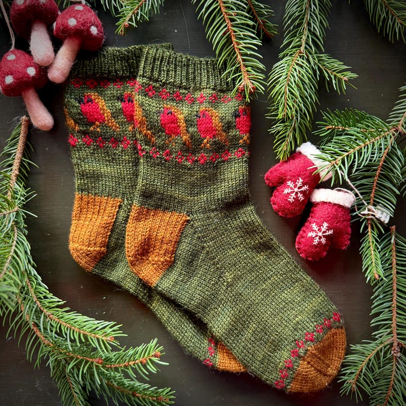 May include: A pair of green knitted socks with a brown and red pattern of robins. The socks have a brown cuff and are lying on a dark brown surface with green pine branches.