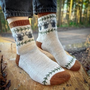 May include: A pair of white socks with brown and green knit patterns. The socks feature a repeating design of small grey animals. The socks are worn on a person's feet.