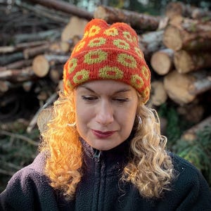 Puede incluir: Una mujer con el pelo rubio rizado lleva una boina de punto naranja y amarilla brillante con un patrón geométrico. La boina tiene un borde doblado y le cubre las orejas. Lleva una chaqueta de vellón negra.