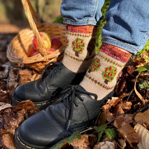 May include: A pair of black leather shoes with laces and yellow stitching, worn with blue jeans and beige socks. The socks feature a green and red tree design. A wicker basket filled with apples is in the background.