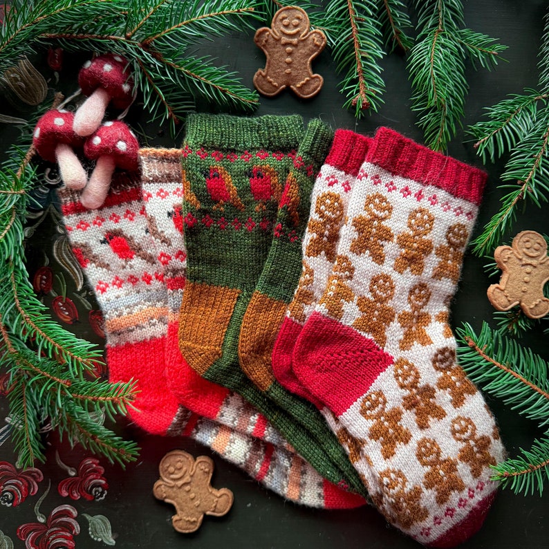 May include: Three pairs of knitted socks with different patterns. The socks are red, green, and white with gingerbread men designs. The socks are arranged on a dark background with a few sprigs of pine needles.