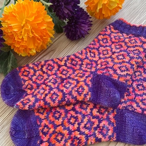 Könnte beinhalten: Ein Paar violette und orangefarbene gestrickte Socken mit geometrischem Muster. Die Socken liegen auf einer Holzoberfläche mit orangefarbenen und violetten Blumen.