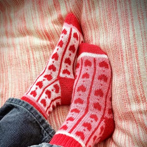 May include: Red and white knitted socks featuring a heart design. The socks are worn with blue jeans and are resting on a striped, reddish-pink fabric. The socks have red heels, toes, and cuffs.