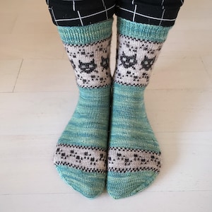 May include: A pair of teal blue and green knit socks with a grey and black cat pattern. The socks have a light grey background with a black cat face pattern.