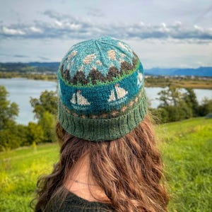 Könnte beinhalten: Eine gestrickte Beanie-Mütze mit nautischem Design. Die Mütze hat eine türkisblaue Krone mit weißen Segelbooten, Bergen und einem grünen Rand. Der Hintergrund zeigt einen See und einen bewölkten Himmel.