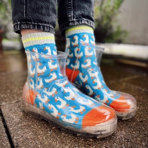 Puede incluir: Botas de lluvia transparentes con un diseño estampado azul y blanco con pequeños patos blancos. Las botas tienen un ribete naranja alrededor de la parte superior.