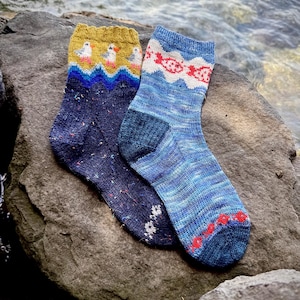 May include: A pair of knit socks. The left sock is dark navy blue with multicoloured flecks and a mustard yellow cuff featuring a pattern of white and orange birds. The right sock is variegated blue with a white wave pattern and red fish motifs.