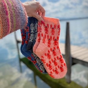 May include: Three pairs of knitted socks are held up against a blurred background of water and sky. The socks are blue, gray, and pink, each patterned with red lobster designs. The socks are made of a soft, knitted material.