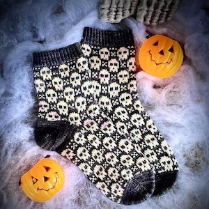 May include: A pair of black and cream-colored knit socks with a repeating skull pattern. The socks have black heels, toes, and cuffs. Two small orange jack-o'-lanterns and a skeleton hand are in the background.
