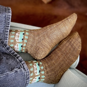 以下が含まれることがあります： 白、黒、ティール色の模様が入った茶色のニットソックス。靴下はリブ編みで、手作りのようです。靴下を履いている人は水色の表面に座っており、青いジーンズが見えます。