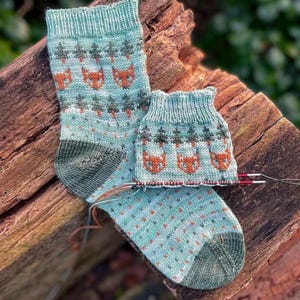 May include: A pair of hand-knitted socks in a light blue yarn featuring a repeating pattern of orange fox faces and dark green pine trees. The socks have solid dark green heels and toes, with a small matching sock cuff on circular knitting needles.