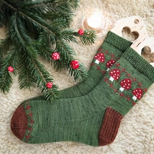 May include: A pair of green knit socks with a pattern of red and white mushrooms. The socks have brown heels and toes. A sprig of evergreen with red berries is in the background.