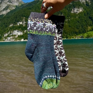 Könnte beinhalten: Ein Paar gestrickte Socken in einer Farbpalette aus Dunkelblau und Anthrazit. Die Socken zeigen ein detailliertes Fair-Isle-Muster mit weißen, hellblauen und grünen Akzenten am Bündchen und an der Spitze in einer strukturierten Strickweise.