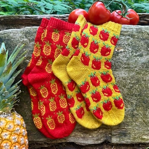 Pode incluir: Dois pares de meias de malha, um vermelho com padrões de abacaxi amarelos e o outro amarelo com padrões de morango vermelhos. Tomates frescos repousam sobre uma superfície de madeira, com um abacaxi à esquerda.