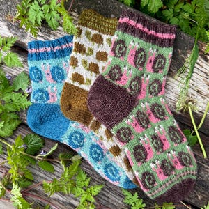 May include: Three pairs of colorful knitted socks with patterned designs. One pair is blue with pink snails, another is brown with white and brown shapes, and the third is green with pink and brown shapes. The socks are displayed on a wooden surface.