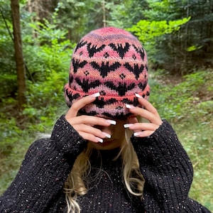 May include: A knitted beanie featuring a repeating pattern of black bat silhouettes against a variegated background of pink, orange, and purple hues. The hat has a folded cuff and is constructed from yarn with a soft, textured appearance.