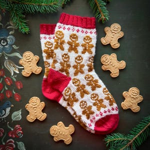 May include: A pair of white and red knit socks with a gingerbread man pattern. The socks are lying on a dark green surface with gingerbread cookies scattered around them.
