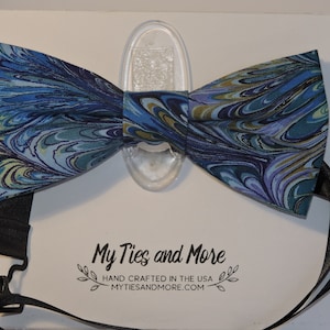 May include: A bow tie with a blue and teal marbled pattern and a black adjustable strap. The bow tie features a swirling design with gold and purple accents. The text "My Ties and More" is visible at the bottom of the image.
