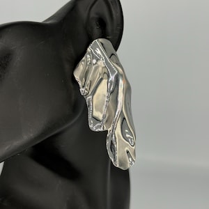 May include: A silver earring with an abstract design. The earring has a textured, flowing shape, reflecting light. The jewelry is displayed on a black mannequin head, showcasing its form and style. The earring's design is modern and eye-catching.
