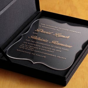 May include: A clear acrylic wedding invitation with gold lettering. The invitation reads "The pleasure of your company is requested by David Lynch and Patricia Ramirez for the celebration of their union on Saturday, August 30, 2018 at Le Chateau d'Esclimont, Rue du Parc, 28700 Bleury-Saint-Symphorien, France. Reception to follow."