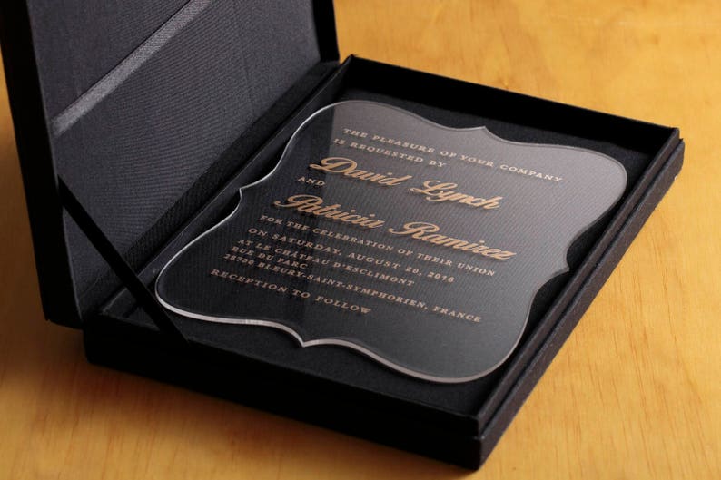 May include: A clear acrylic wedding invitation with gold lettering. The invitation reads "The pleasure of your company is requested by David Lynch and Patricia Ramirez for the celebration of their union on Saturday, August 30, 2018 at Le Chateau d'Esclimont, Rue du Parc, 28700 Bleury-Saint-Symphorien, France. Reception to follow."