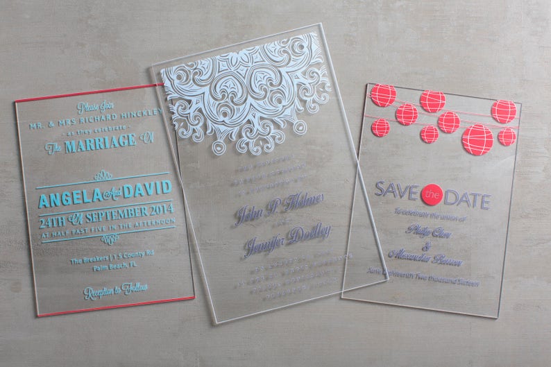 May include: Three clear acrylic wedding invitations with different designs. The first invitation has a white lace design and the text "The Marriage of Angela and David". The second invitation has the text "Helen P. Fisher and Jennifer Dollley". The third invitation has a red and white string of paper lanterns design and the text "Save the Date".