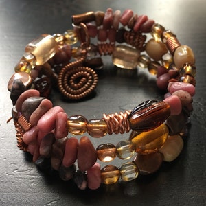 May include: A multi-strand bracelet with copper wire, brown, pink, and clear beads. The bracelet is wrapped around a dark surface.