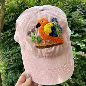 May include: A light pink baseball cap with an embroidered parrot design. The parrot is orange, yellow, and blue, perched on a brown branch with green leaves and purple flowers. The cap has a curved brim and a button on top.