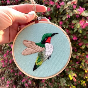 May include: A hand-stitched embroidery hoop featuring a hummingbird with a green, white, and brown body, a red throat, and a black beak. The hoop is on a light blue background.