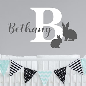 May include: White letter B with the name Bethany in cursive script above it. Two grey bunny silhouettes are to the right of the letter. A white cot with a mint green and black patterned bunting is in the foreground.