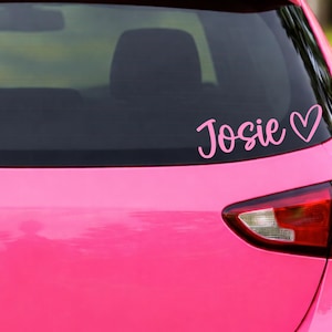 Könnte beinhalten: Ein rosa Vinyl-Aufkleber auf der Heckscheibe eines leuchtend rosa Autos. Der Aufkleber zeigt den Namen Josie in einer geschwungenen Schreibschrift, gefolgt von einem einfachen Herzsymbol. Der Text und das Herz sind in einem hellen Rosaton.