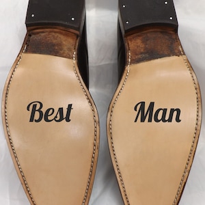 May include: Two brown leather shoes with black soles. The left shoe has the word "Best" written on the sole. The right shoe has the word "Man" written on the sole.