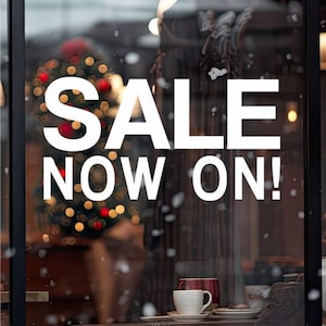 Op de afbeelding: Een wit bord met de tekst "SALE NOW ON!" in een winkelruit. De etalage is versierd met een kerstboom en sneeuw-effecten.