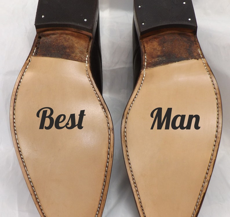 May include: Two brown leather shoes with black soles. The left shoe has the word "Best" written on the sole. The right shoe has the word "Man" written on the sole.