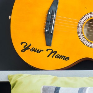 May include: A yellow acoustic guitar with black strings and a black pickguard. The guitar has a decorative design around the sound hole and the words "Your Name" written in black cursive on the body.