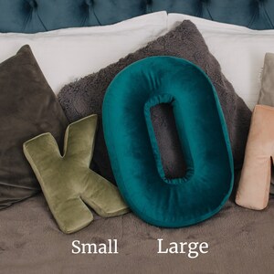 May include: A plush velvet throw pillow shaped like the letter K in olive green and a plush velvet throw pillow shaped like the letter O in teal. The text Small is positioned below the K pillow and the text Large is below the O pillow.
