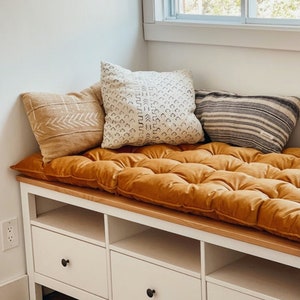 May include: A white wooden bench with three drawers and open storage compartments, topped with a long, tufted mustard yellow cushion. Three decorative throw pillows in tan, cream with black geometric patterns, and grey stripes rest on top.